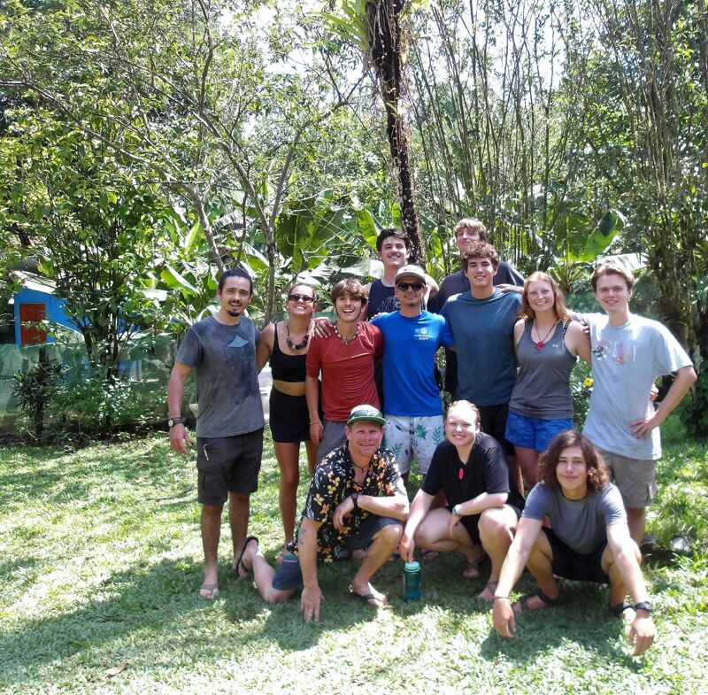 A group of about a dozen people are gathered outside in a grassy, wooded area. They appear to be posing for a photo. Most of them are standing, while a few are kneeling or crouching in the front. The group consists of both men and women, and they are dressed in casual clothing. The background features lush greenery, including trees and various plants.